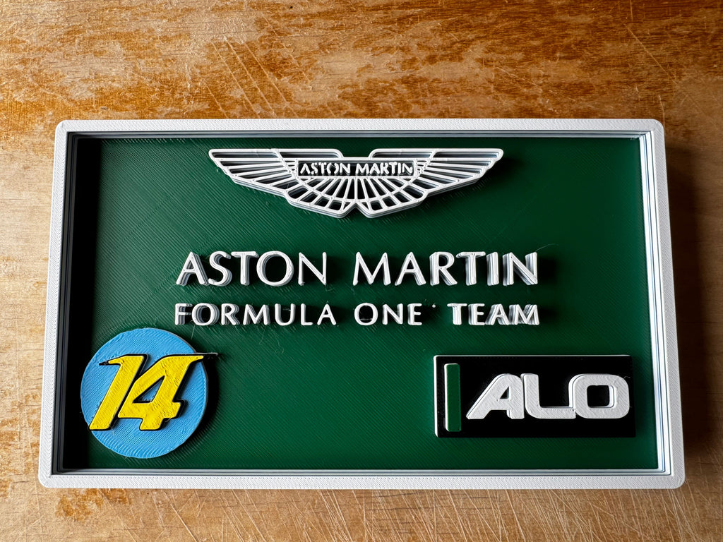 A framed Aston Martin Formula One Team display plaque on a wooden surface. The plaque features a dark green background with raised silver lettering reading 'ASTON MARTIN FORMULA ONE TEAM', a blue and yellow circular '14' emblem, a silver Aston Martin wing logo, and a black-and-silver 'ALO' badge.